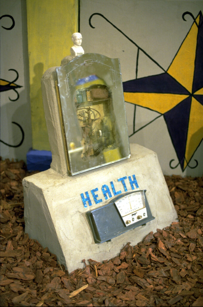 Health, a concrete grave monument from Radio Ancestrale, a mirrored medicine cabinet above the title and a teal radio, a small ceramic head at the peak