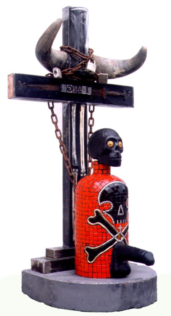 Bizango Altar with Ghede Libation Bottle, a red tile mosaic bottle with a black skull and crossbones and a ceramic skull stopper, standing on a round concrete base from which a steel cross rises, hung with chains, animal horns, and a padlock