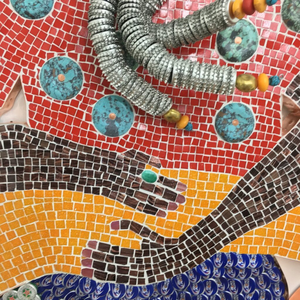 Detail of La Siren III, three materials meeting at once: red glass tesserae set with discs of blue-green dyed stone, silver bottle caps stacked into a coil of hair, and dark brown glass hands laid over orange glass, with a small green stone set into the back of one hand