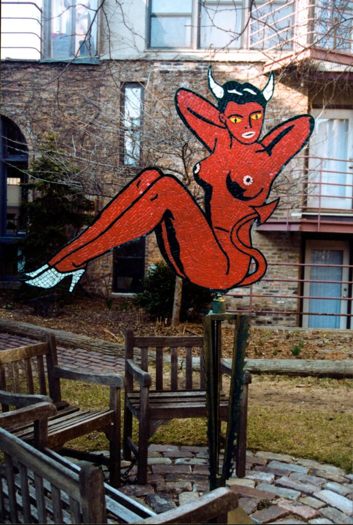 Pomba Gira, a mosaic wind vane in red ceramic tile, a horned nude figure reclining in a pin-up pose with white heels and yellow eyes, mounted on a post in a brick courtyard